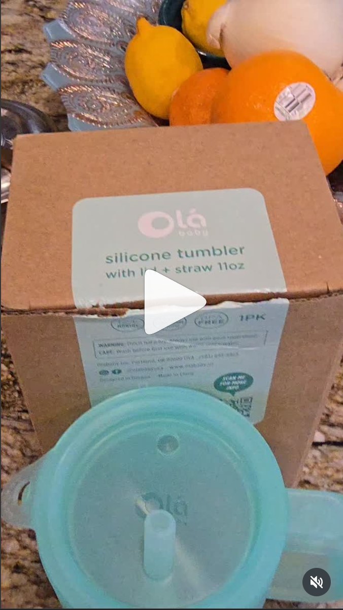 Video still: an Olababy box printed "silicone tumbler with lid + straw 11oz" stands on a kitchen counter beside lemons and oranges, with the tumbler's teal lid and straw in front of it.