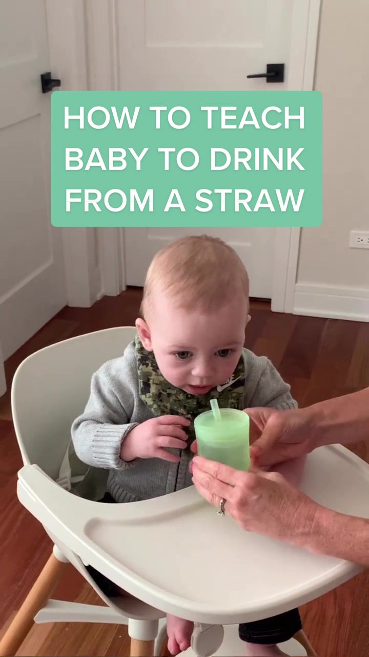 Video still: a baby sits in a white highchair holding a green Olababy training cup with a straw while an adult hand steadies it. Overlay text reads "How to teach baby to drink from a straw".