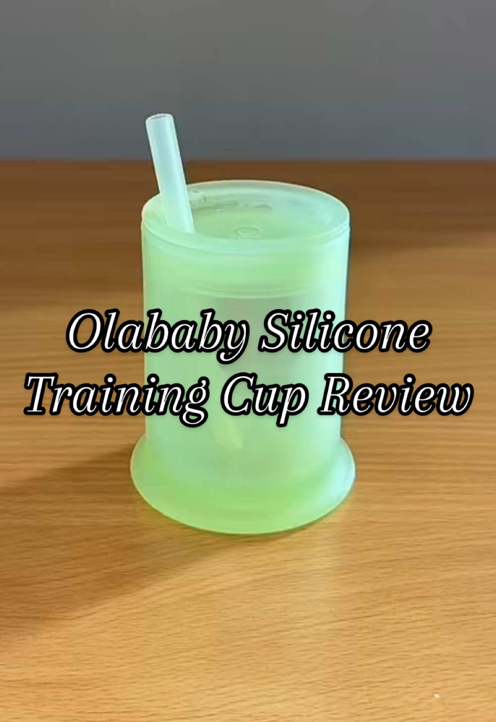 Video still: a green Olababy silicone training cup with a straw in the lid stands on a wooden table. Overlay text reads "Olababy Silicone Training Cup Review".