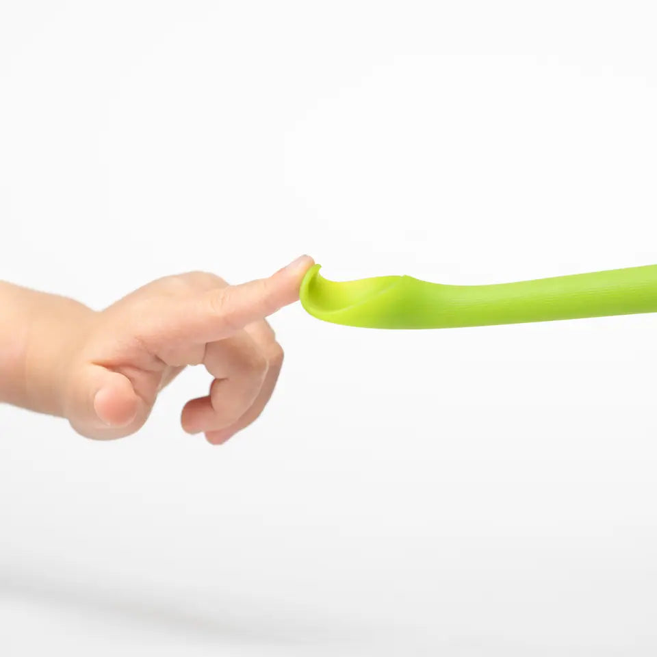 A finger pressing the flexible tip of a green Olababy feeding spoon flat, photographed on white.