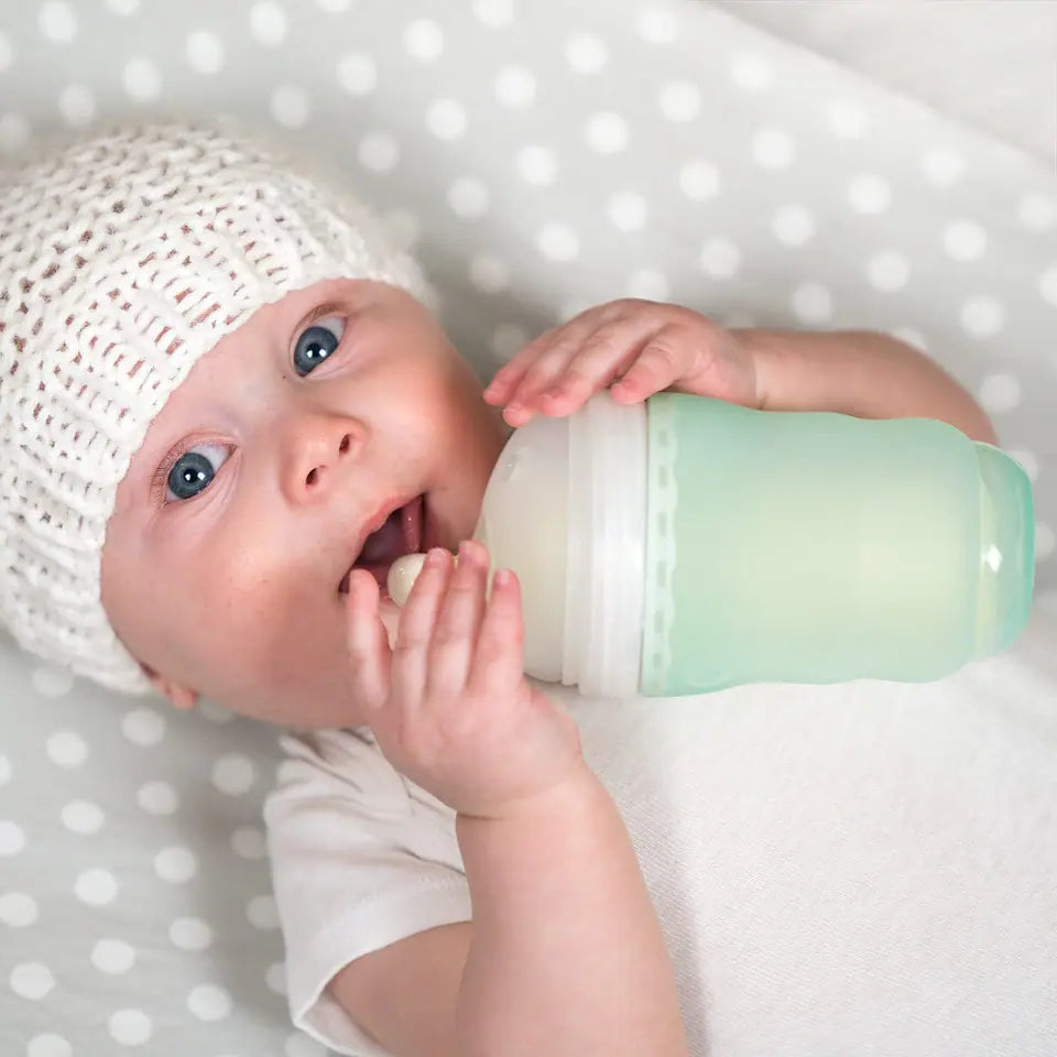 A newborn in a knitted bonnet lying on a spotted blanket, one hand resting on a mint silicone GentleBottle while feeding from it