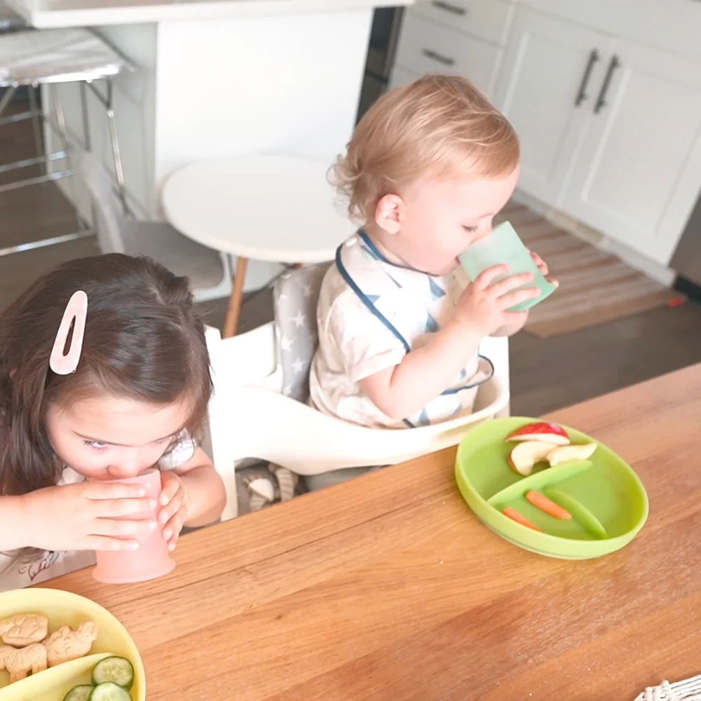 A toddler in a high chair drinking from a mint silicone cup, an older girl at the same table holding a coral silicone cup in both hands, and a green silicone plate of apple slices and carrot sticks between them